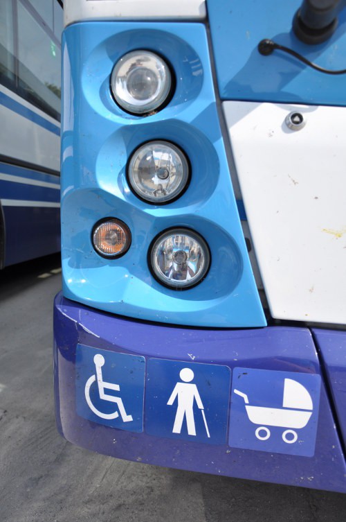 Prezentacja biało-niebieskiego autobusu Solaris - Zbliżenie na prawe światła drogowe, pod nimi znajdują się trzy niebieskie naklejki informujące, że pojazd jest dostosowany do osób poruszających się na wózku inwalidzkim, dla niewidomych oraz wózków dziecięcych.