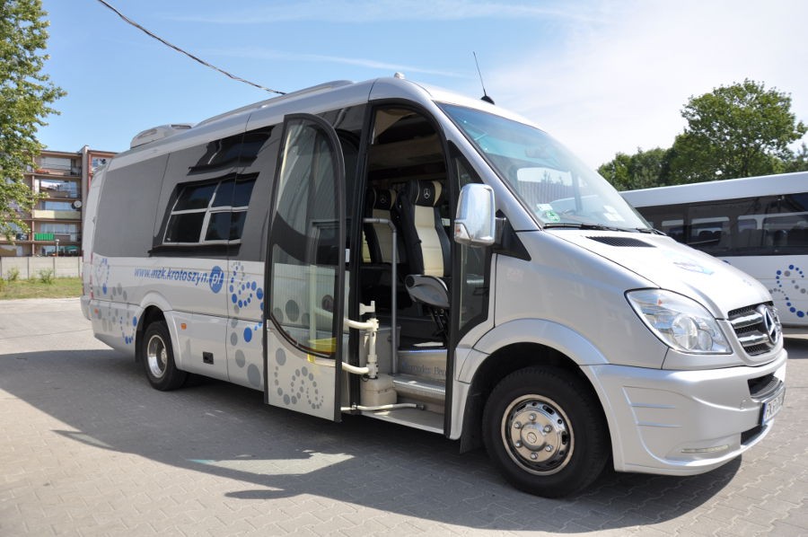 Prezentacja busa Mercedes, pojazd ustawiony prawym bokiem, otwarte drzwi. Widoczne niebieskie logo MZK w Krotoszynie wraz z niebieskim adresem strony internetowej.