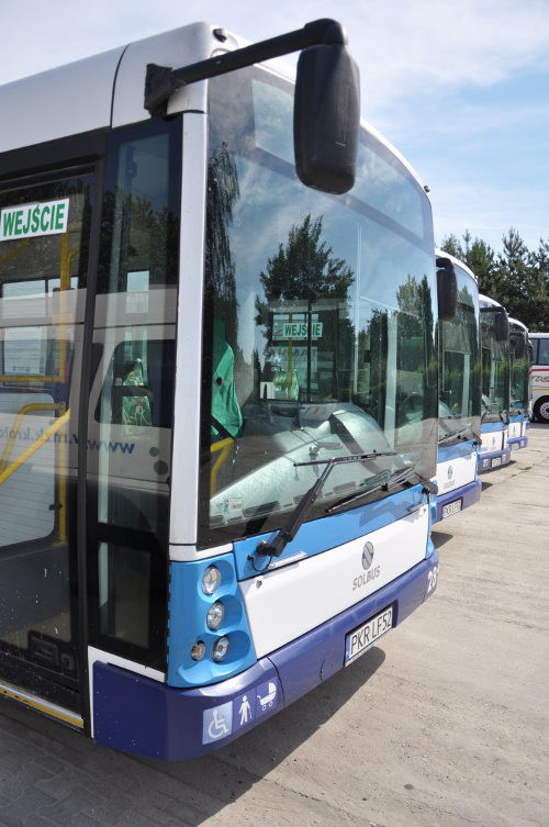 Prezentacja biało-niebieskiego autobusu Solaris - widoczny przód oraz prawy bok pojazdu. Zbliżenie na prawe lusterko kierowcy oraz drzwi. W tle ustawione w rzędzie inne 4 takie same modele autobusów.